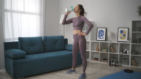 sporty-woman-is-standing-in-living-room-and-relaxing-drinking-water-break-at-morning-workout-at-home-fitness-and-wellness